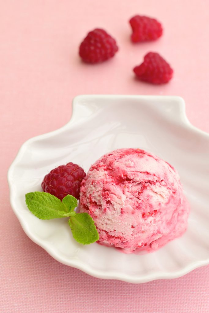 Eine Kugel Himbeereis mit Himbeere und Minze liegt auf einem weißen Teller.