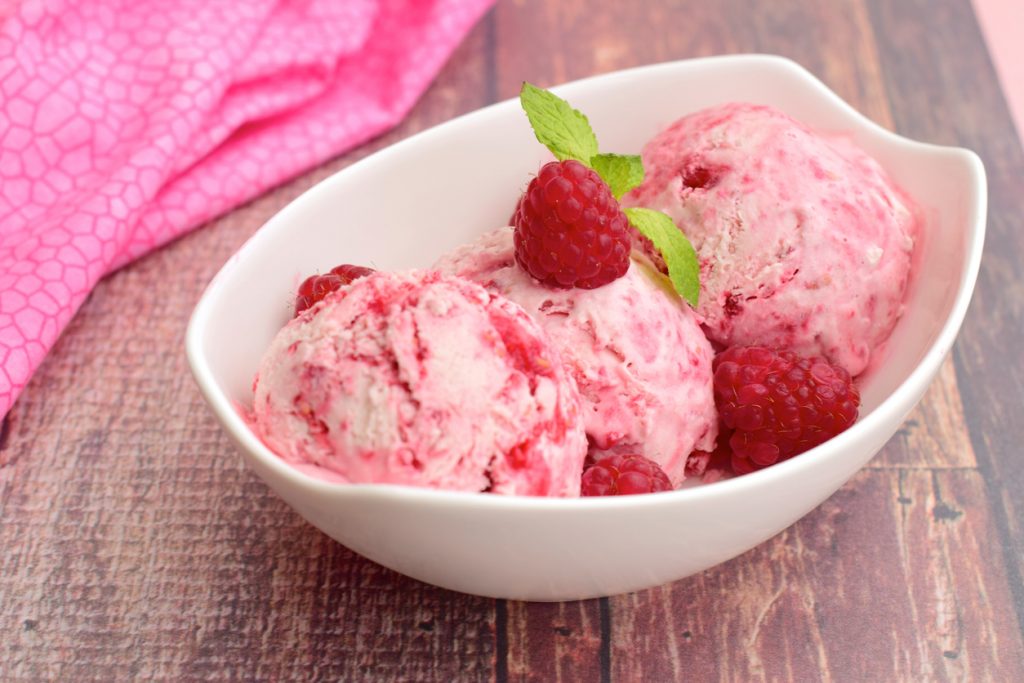 Drei Kugeln Himbeereis liegen mit frischen Himbeeren in einer weißen Schale.