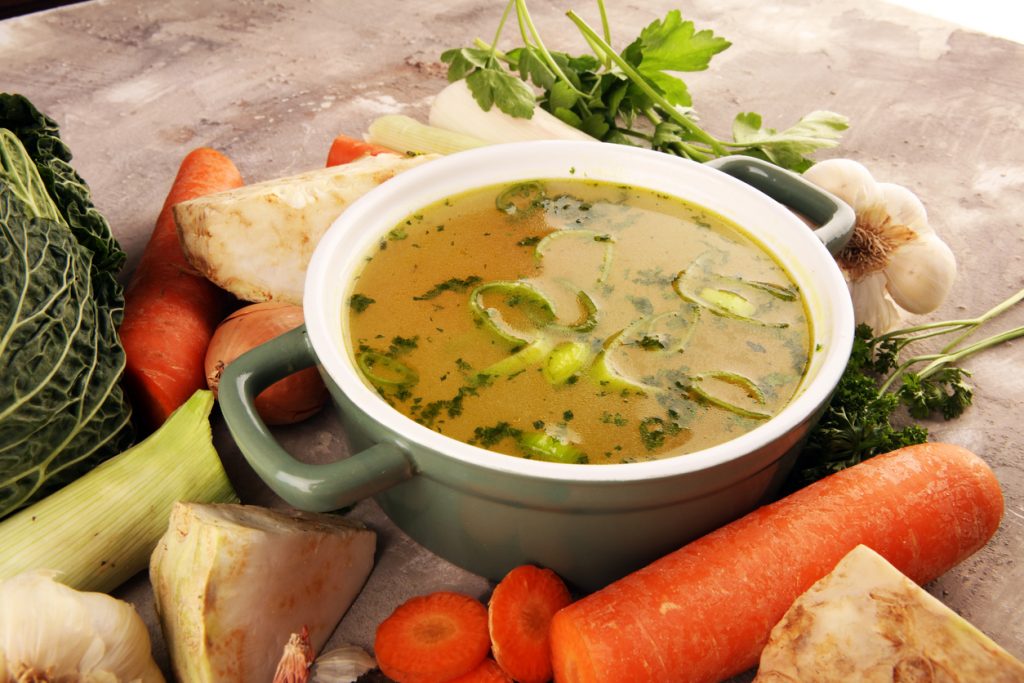 Eine Schüssel Gemüsebrühe steht in einem Topf, umgeben von Karotten, Lauch, Knollensellerie und Kräutern.