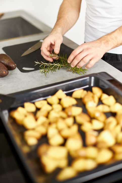 Hände zupfen frische Rosmarinzweige über einem Backblech mit rohen Kartoffelwürfeln ab.