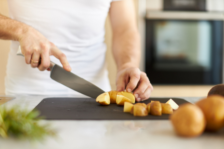 Ein Mann schneidet auf einem dunklen Brett Kartoffeln in gleichmäßige Würfel, im Hintergrund ist ein Backofen zu sehen.