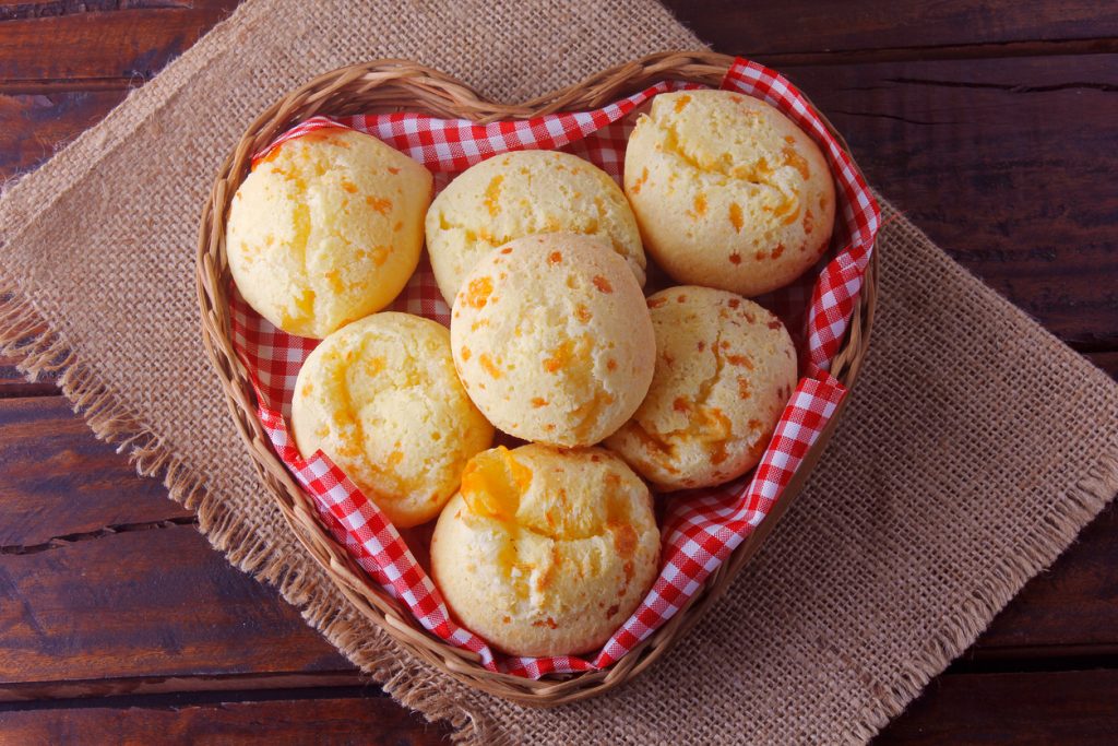 Acht Pão de Queijo liegen in einem herzförmigen Körbchen, das mit einem karierten Tuch ausgelegt ist.