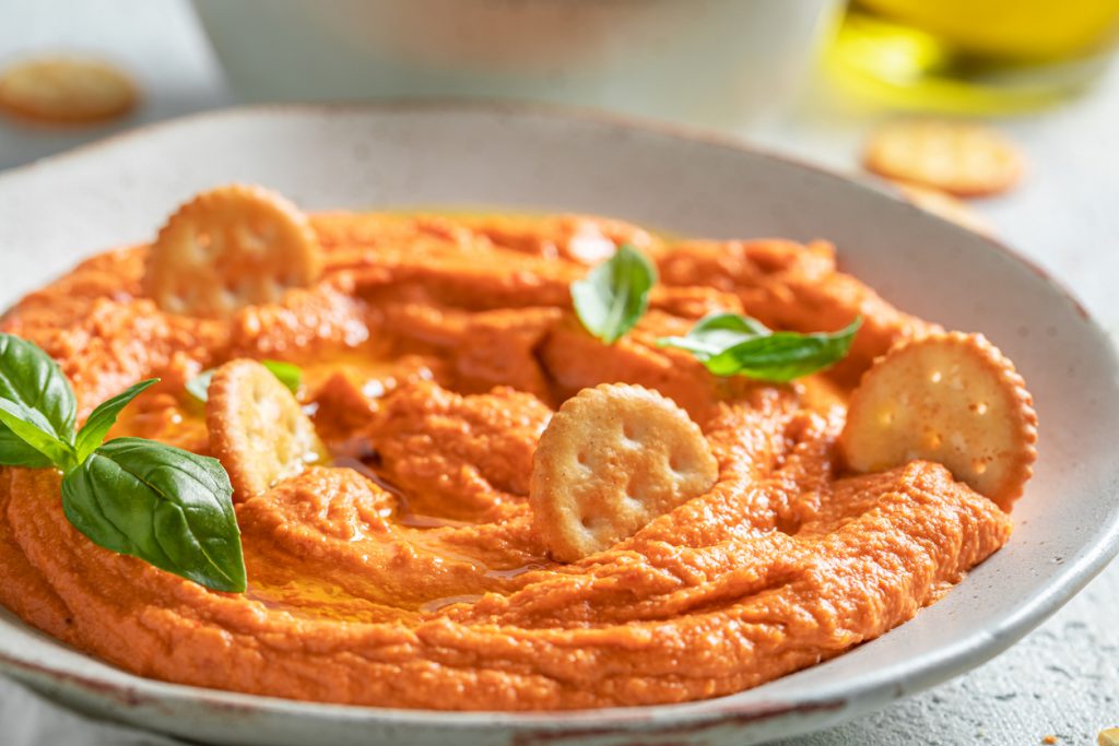 Eine Nahaufnahme zeigt die feine Struktur des Tomatenhummus in einer Schale mit eingetauchten Crackern und glänzendem Olivenöl.