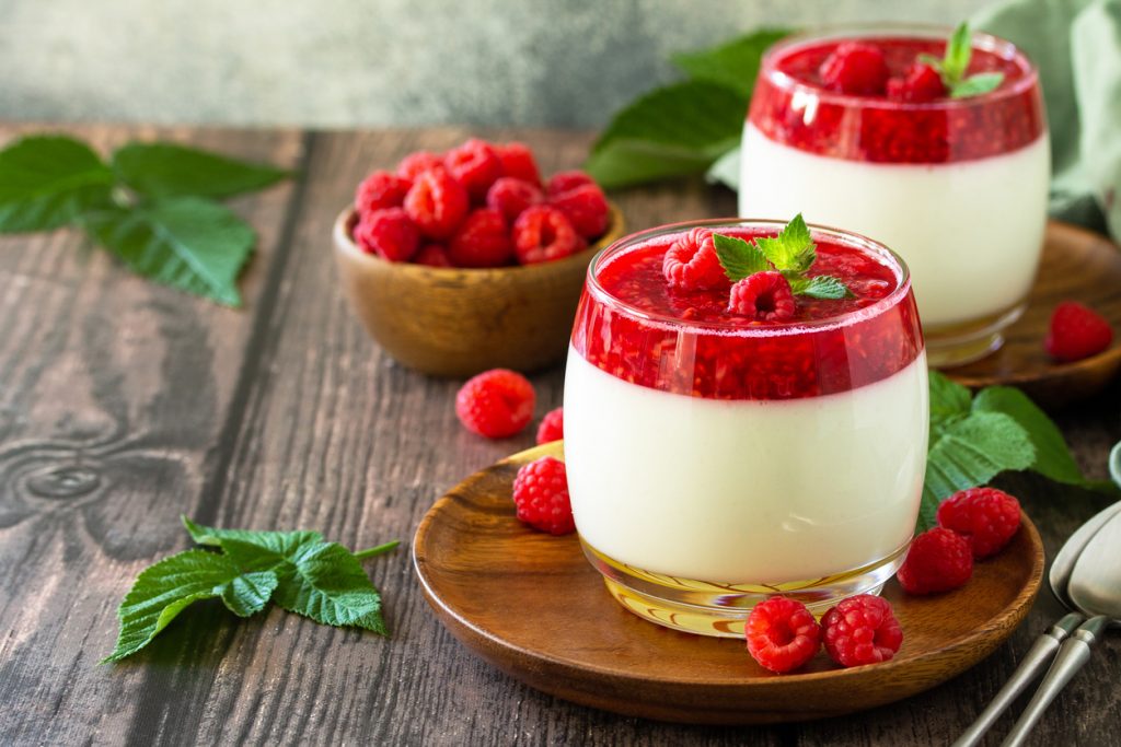 Ein Glas Panna Cotta mit Himbeerpüree steht im Fokus auf einem dunklen Holztisch.