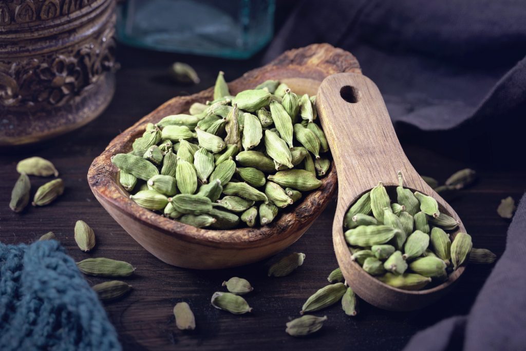 Ganze Kardamomkapseln liegen in einer Holzschale und auf einem hölzernen Löffel.