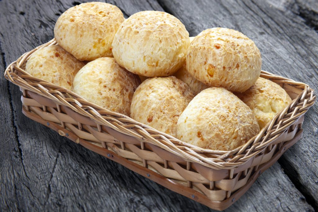 Ein geflochtener Korb ist bis oben hin mit runden Pão de Queijo gefüllt und steht auf einer dunklen Holzoberfläche.