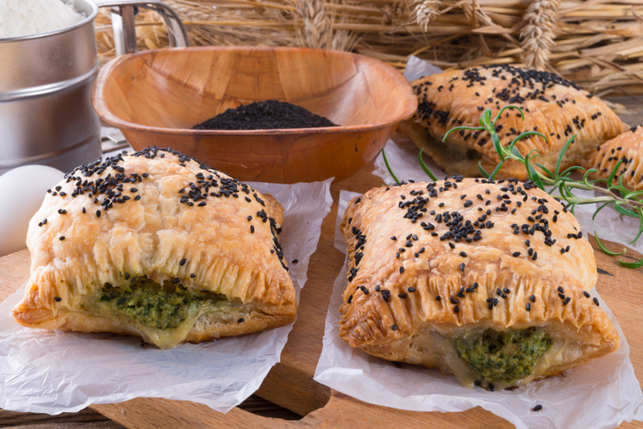 Zwei goldbraun gebackene Blätterteigtaschen mit Spinatfüllung und Schwarzkümmel-Bestreuung liegen auf weißem Backpapier.