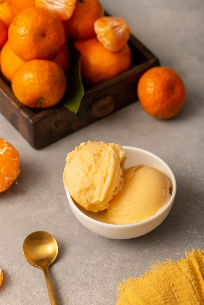 Hochformatige Ansicht von zwei Kugeln Mandarineneis in einer weißen Schale neben einem goldenen Löffel und einer Mandarinenkiste.