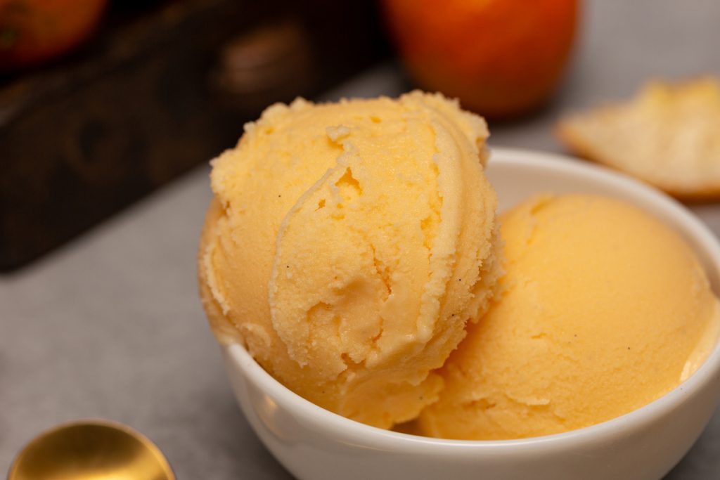 Nahaufnahme von zwei cremigen Kugeln Mandarineneis in einer weißen Keramikschale auf grauem Hintergrund.