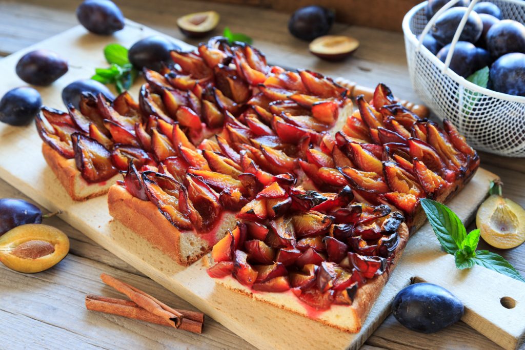 Angeschnittener Pflaumenhefekuchen steht auf einem Holzbrett, mit frischen Pflaumen im Hintergrund.