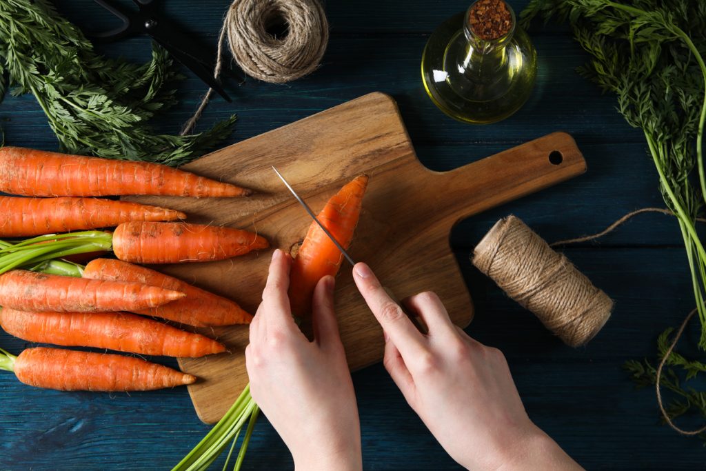 Eine Person schneidet Möhren auf einem Holzbrett, umgeben von frischen Karotten und Küchenutensilien.