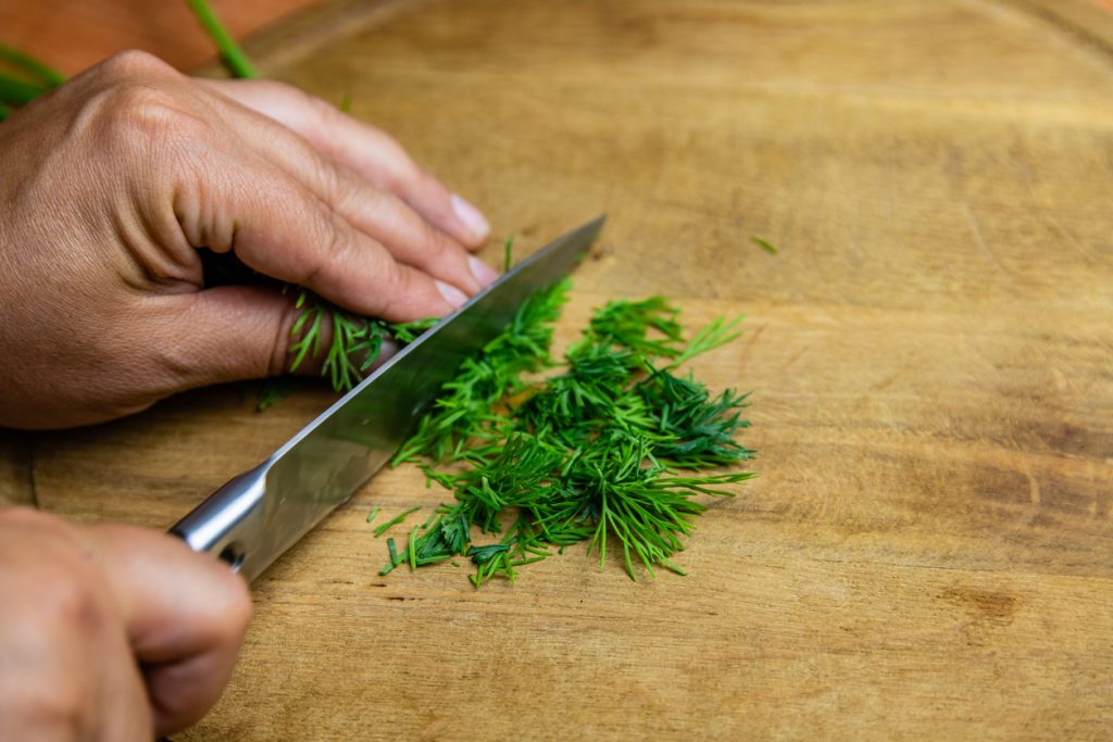 Hände schneiden frischen grünen Dill mit einem scharfen Messer auf einem runden Schneidebrett aus Holz.