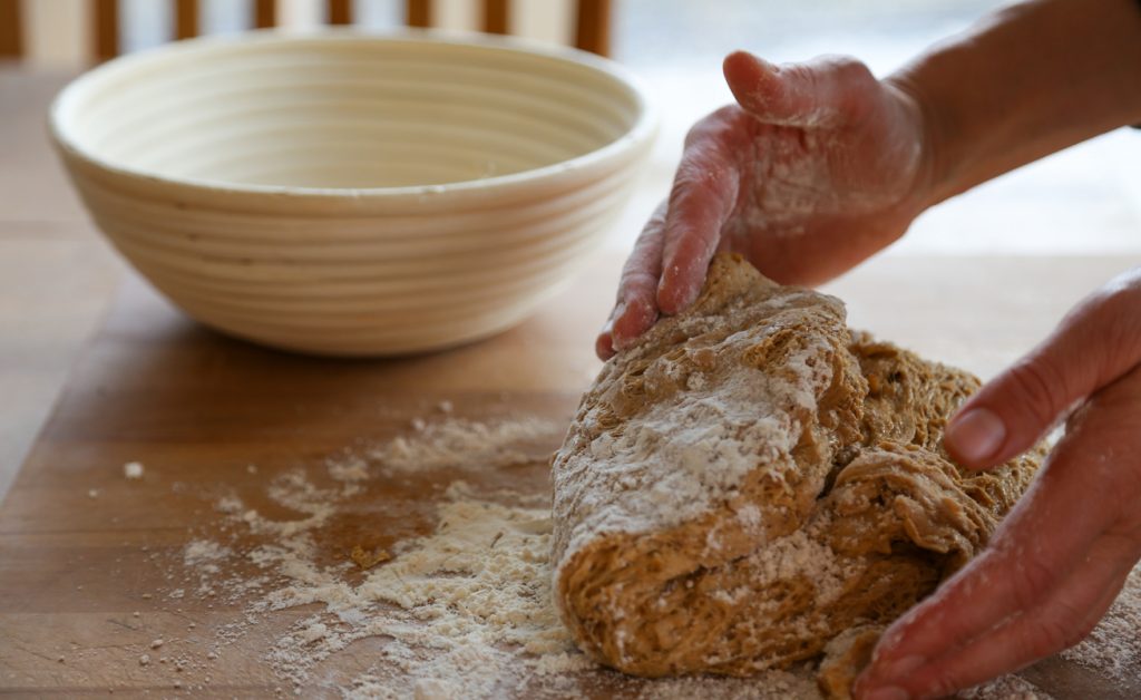 Mehlige Hände kneten einen dunklen Roggenteig auf einer Holzfläche neben einem Gärkörbchen.