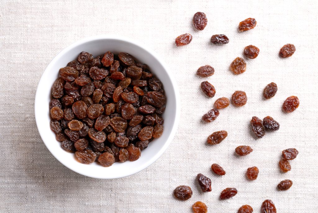 Eine weiße Schale mit Rosinen liegt auf hellem Stoff, daneben verstreute Rosinen.