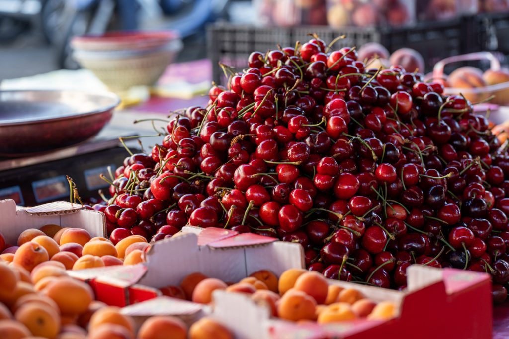 Ein großer Haufen tiefroter, glänzender Kirschen mit grünen Stielen liegt auf einem Markttisch neben Kisten mit Aprikosen.