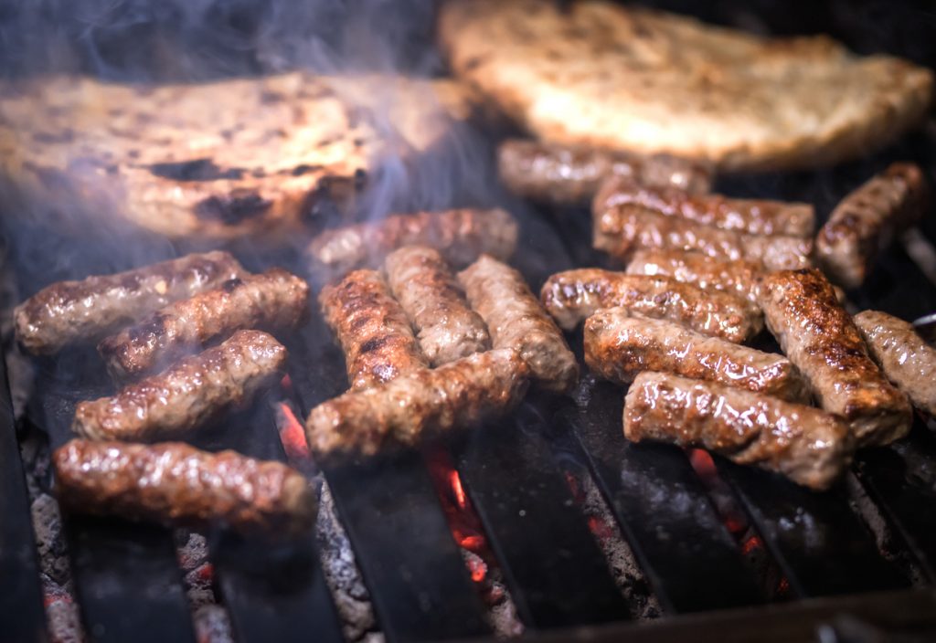 Mehrere Cevapcici liegen auf einem heißen Grillrost mit glühender Kohle und Rauch.