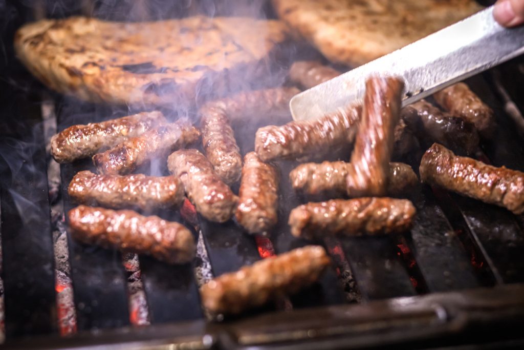 Eine Zange wendet Cevapcici auf dem Grill, während im Hintergrund Fladenbrot mitröstet.