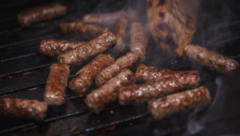 Cevapcici liegen saftig auf dem Grillrost, im Hintergrund ist ein Stück Fladenbrot zu sehen.