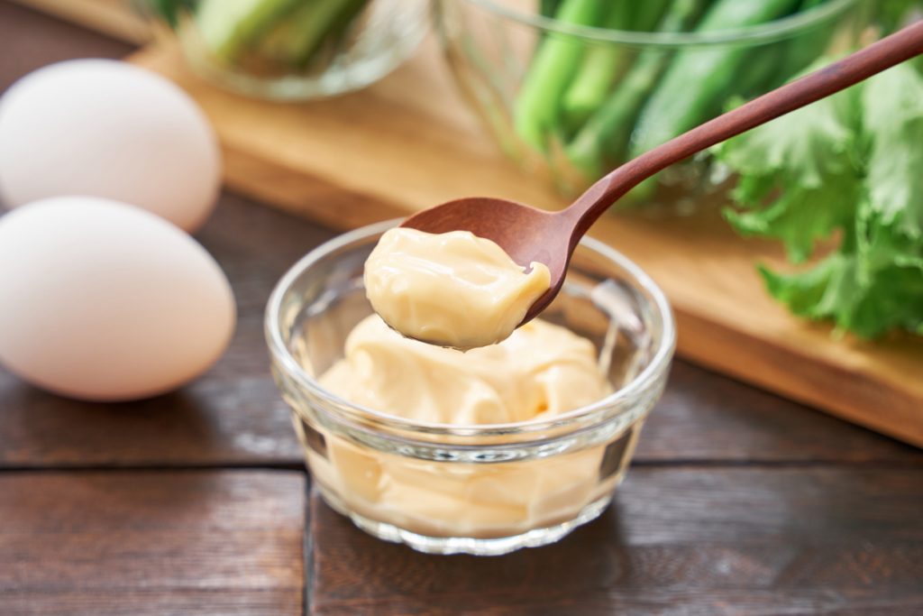 Ein Holzlöffel hebt eine Portion cremige Mayonnaise aus einer kleinen Glasschale.