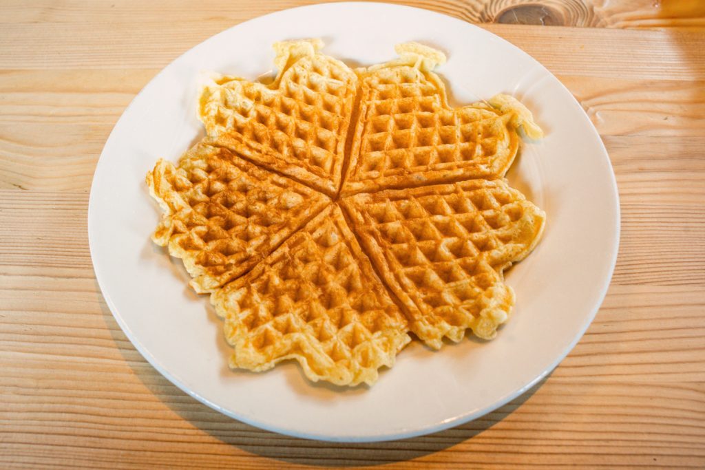 Eine große, runde Waffel, die aus fünf zusammenhängenden Herzen besteht, liegt flach auf einem weißen Teller auf einem Holztisch.