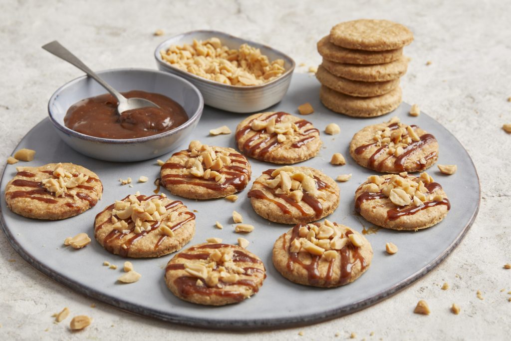 Haferkekse mit Karamell und Topping neben einer Schüssel Karamellsauce und gestapelten Keksen.