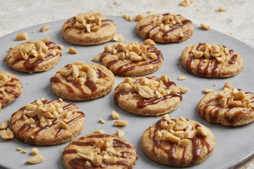 Haferkekse mit Karamellstreifen und gehackten Nüssen auf einem Blech angerichtet.