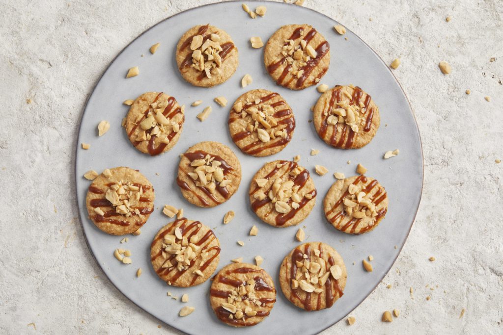 Runde Haferkekse mit Karamellstreifen und gehackten Nüssen auf einem großen Teller angerichtet.