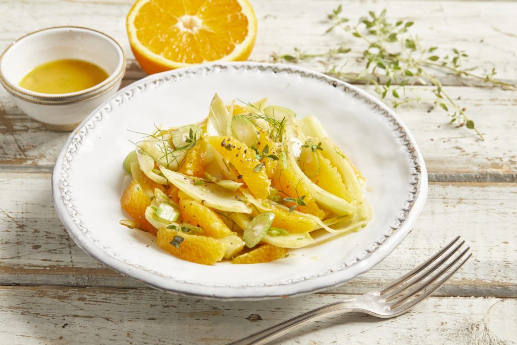 Fenchel-Orangen-Salat liegt auf einem weißen Teller mit Kräutern, im Hintergrund sind Orange und Dressing zu sehen.