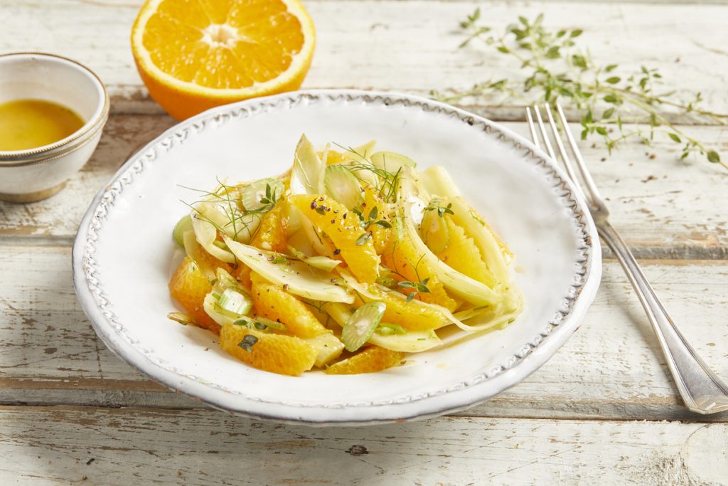 Fenchel-Orangen-Salat liegt auf einem weißen Teller, daneben liegen Besteck, Kräuter und eine halbe Orange.