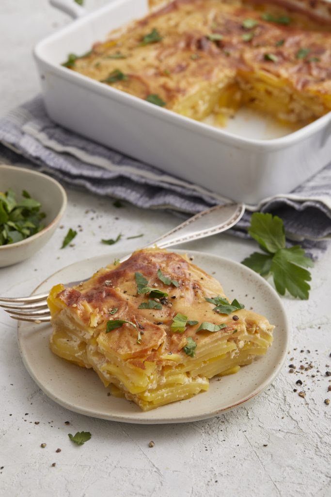 Ein Stück Kartoffel-Gratin liegt auf einem Teller und ist mit Petersilie und Pfeffer bestreut.