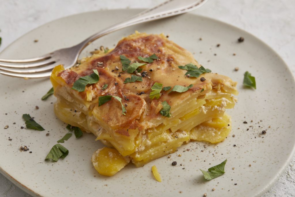 Ein Stück Kartoffel-Gratin liegt auf einem hellen Teller neben einer Gabel und ist mit Kräutern garniert.