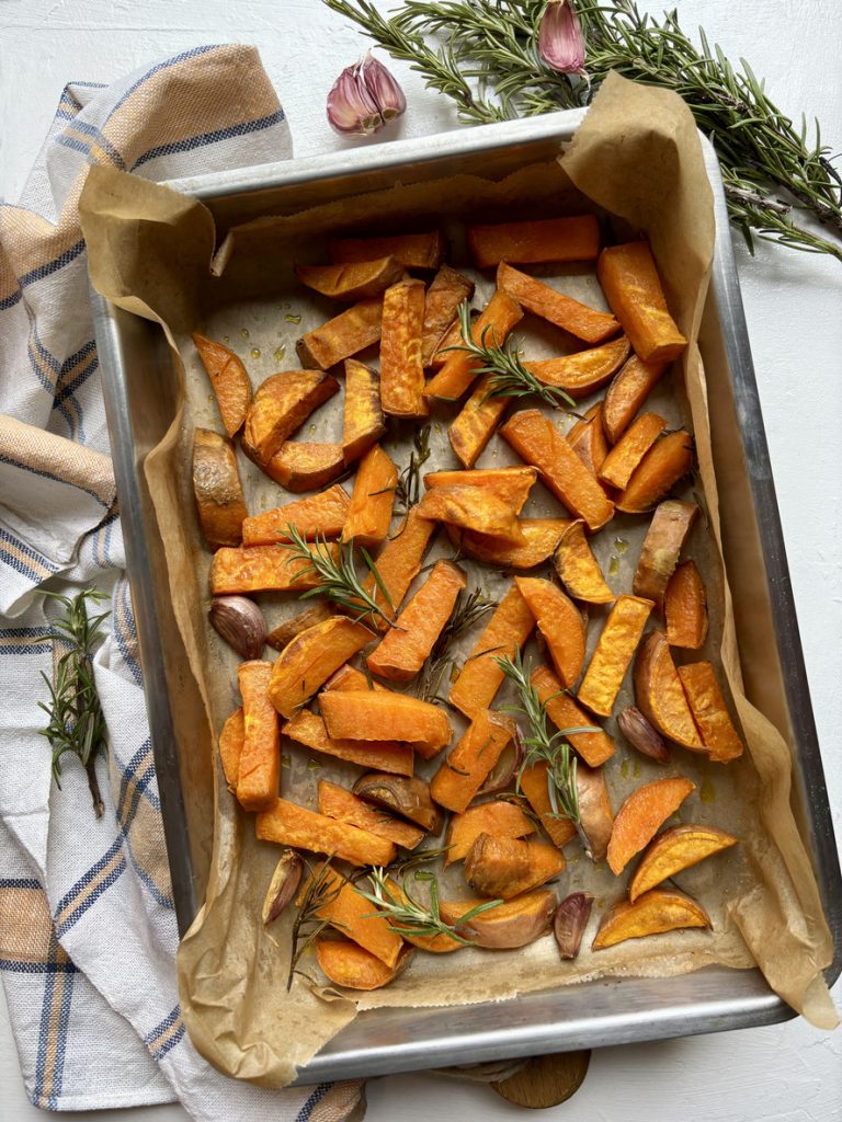 Goldbraun gebackene Süßkartoffelstücke liegen mit frischen Rosmarinzweigen auf Backpapier.