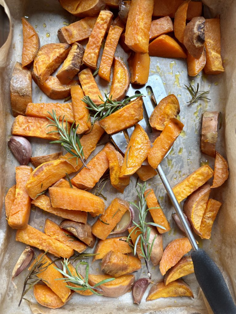 Eine Backform mit Süßkartoffeln steht neben einem Küchentuch und frischen Kräutern auf hellem Untergrund.