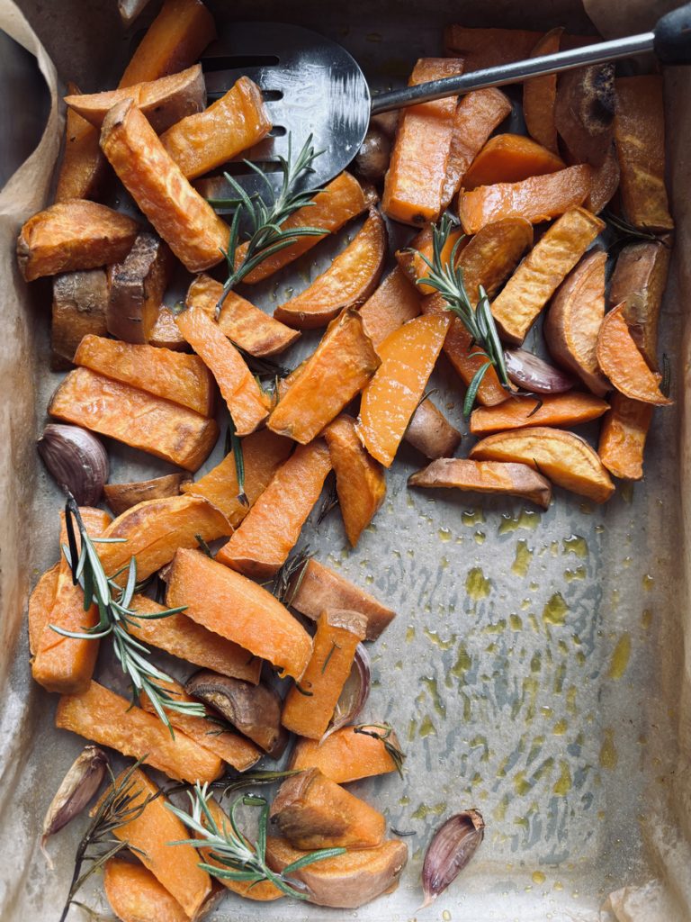 Ein metallisches Backform-Tablett ist mit knusprigen Süßkartoffelspalten und Knoblauch gefüllt.