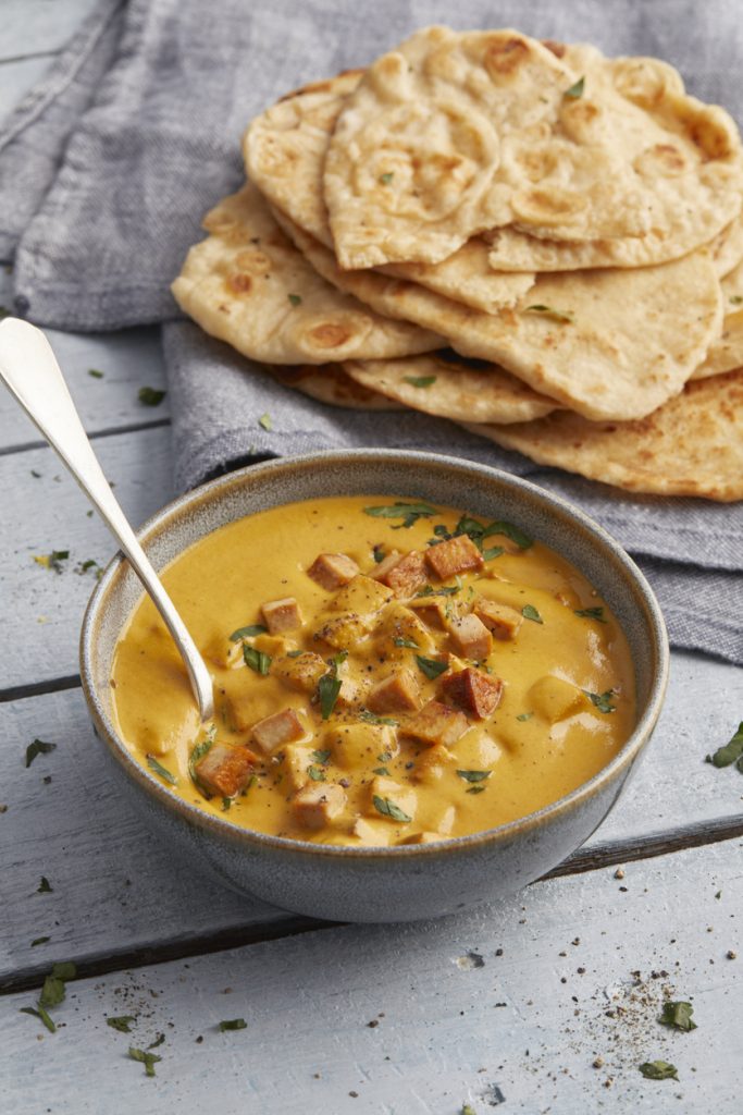 Eine Schale mit indischem Tofu Tikka Masala, in der ein silberner Löffel steckt, daneben liegt Naan-Brot auf einem grauen Tuch.