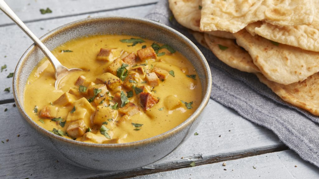 Eine Schüssel cremiges Tofu Tikka Masala auf einem blauen Holztisch, serviert mit einem Stapel Naan-Brot im Hintergrund.