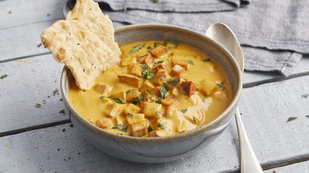 Nahaufnahme einer Schüssel Tofu Tikka Masala, in die zwei Stücke Fladenbrot eingetaucht sind, auf einem hellblauen Holztisch.