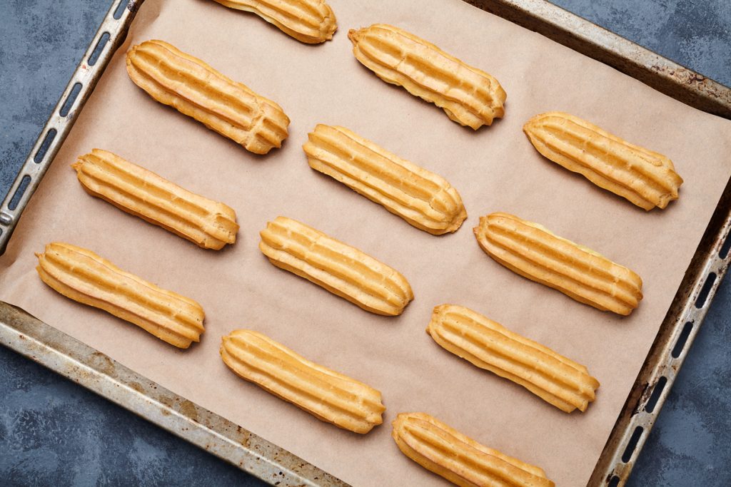 Ein Backblech mit Pergamentpapier, auf dem zwölf gleichmäßig gebackene, längliche Brandteig-Stangen in Reihen angeordnet sind.