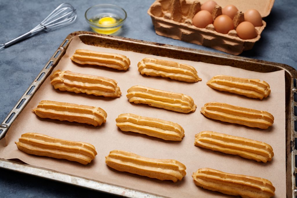 Ein volles Backblech mit Eclairs im Vordergrund; dahinter stehen eine Schale mit einem aufgeschlagenen Ei, ein Schneebesen und ein Karton mit braunen Eiern.