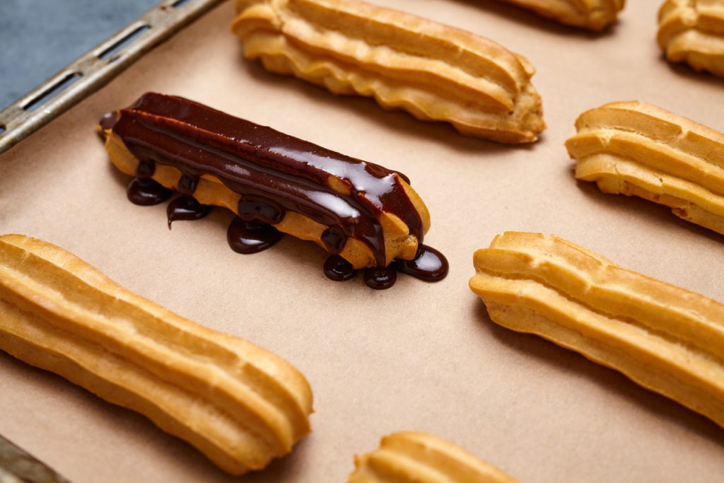 Nahaufnahme eines Eclairs auf Backpapier, das gerade mit einer dicken, glänzenden Schicht dunkler Schokoladenglasur überzogen wurde.