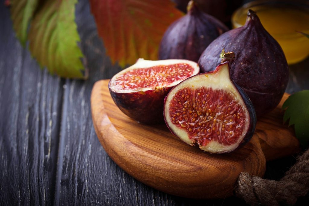 Zwei halbierte, tiefrote Feigen liegen neben ganzen violetten Früchten auf einem Holzbrett vor dunklem Hintergrund.