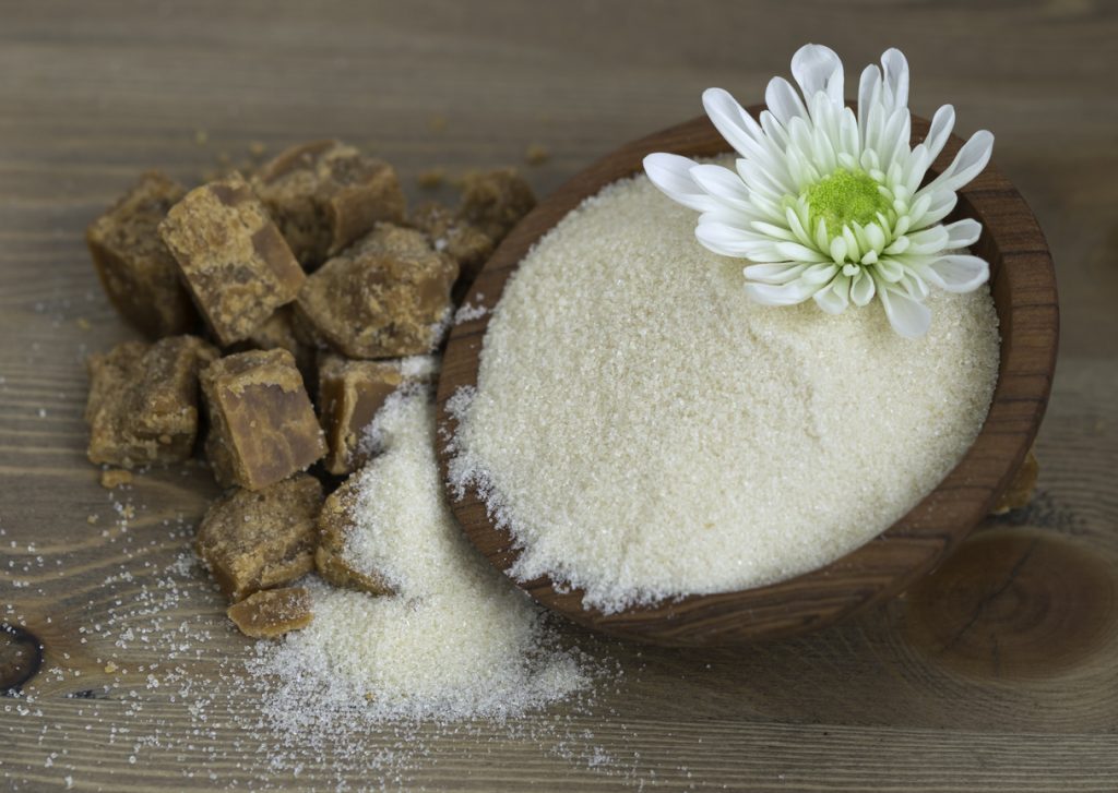 Kokosblütenzucker liegt in einer Holzschale, daneben liegen braune Zuckerbrocken und etwas verstreuter Zucker.