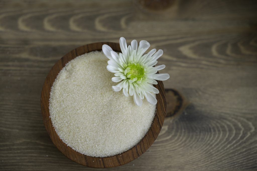 Eine Holzschale voller Kokosblütenzucker steht auf einem Holztisch und ist mit einer weißen Blüte dekoriert.