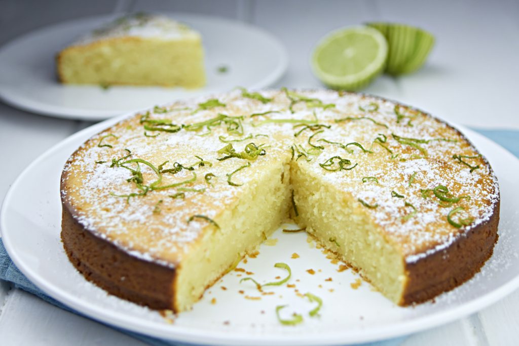 Ein angeschnittener, runder Kuchen auf einem weißen Teller, bestreut mit Puderzucker und frischen Limettenzesten; im Hintergrund ist ein weiteres Kuchenstück und eine aufgeschnittene Limette zu sehen.