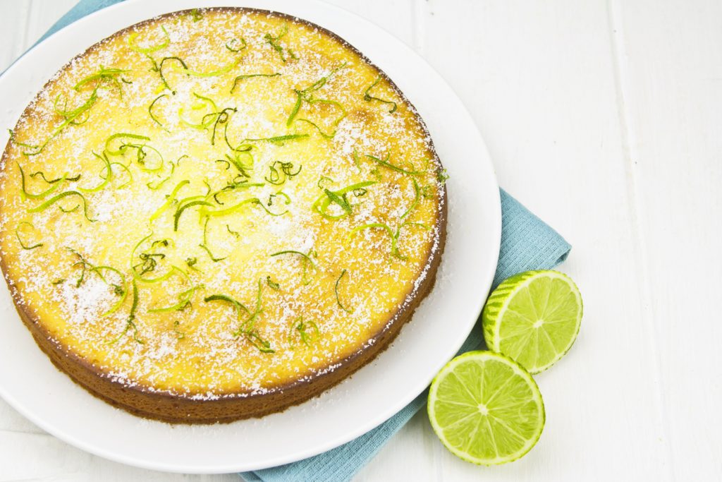 Die Ansicht von oben zeigt den gesamten runden Kuchen auf einer weißen Platte, großzügig mit Puderzucker und spiralförmigen grünen Limettenzesten garniert.