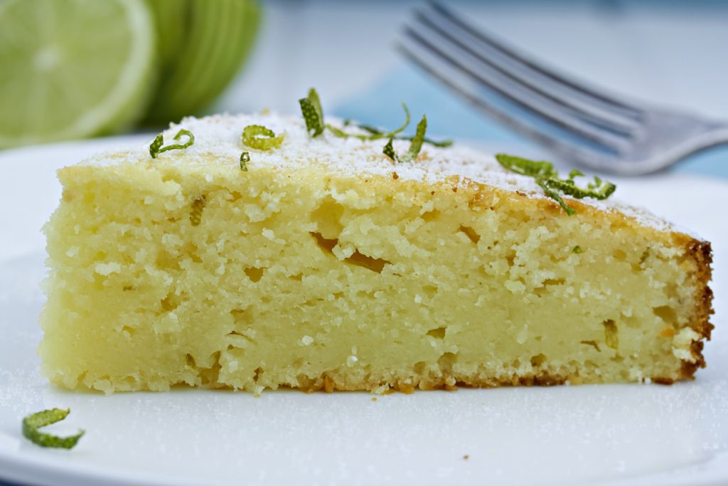 Die Makroaufnahme eines einzelnen Kuchenschnitts auf einem weißen Teller zeigt die feuchte Krume und die leichte Kruste am Rand, garniert mit Limettenabrieb.