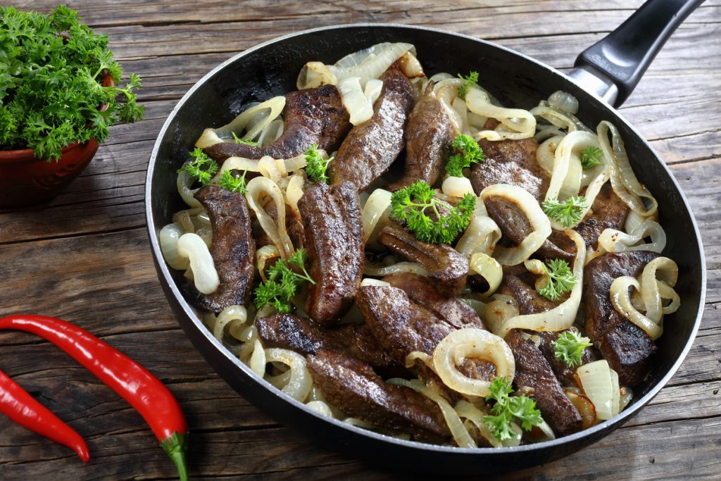 Eine Pfanne mit gebratener Kalbsleber und Zwiebelringen steht auf einem rustikalen Holztisch neben Kräutern.