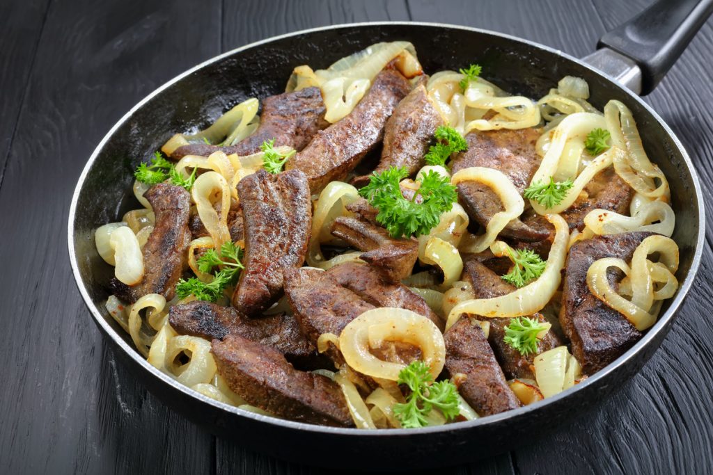 Gebratene Kalbsleberstücke sind mit Zwiebelringen und Petersilie in einer Pfanne angerichtet.