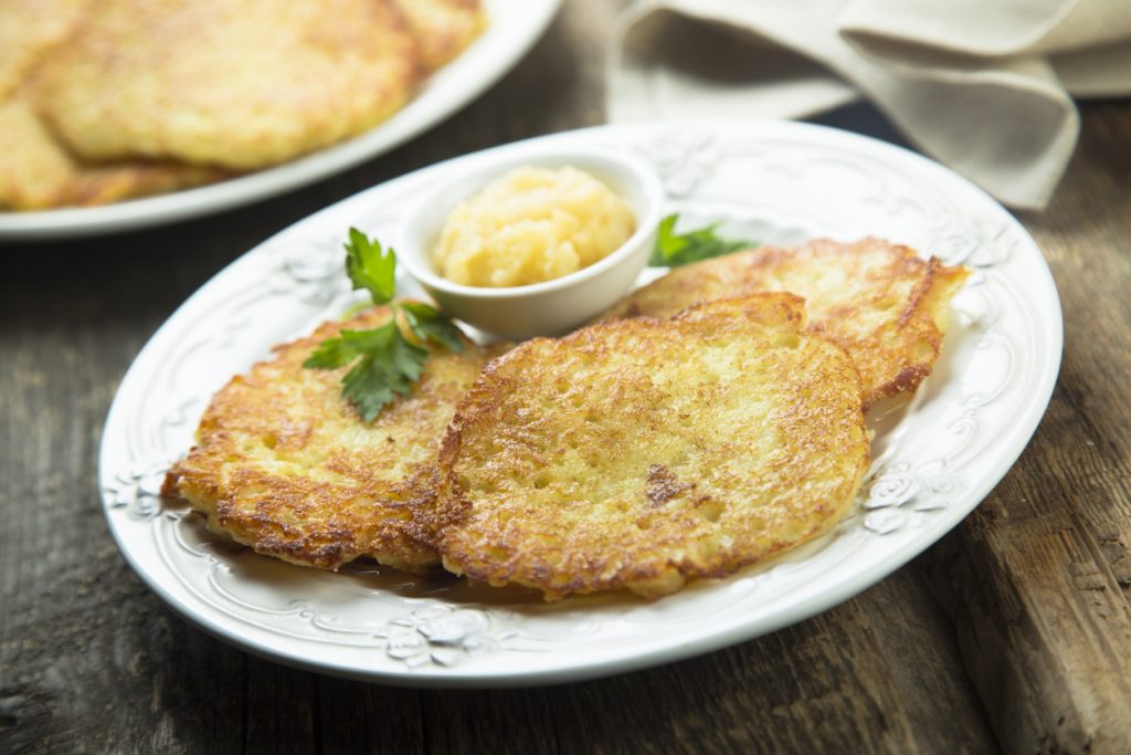 Zwei Kartoffelpuffer liegen auf einem weißen Teller und werden mit Apfelmus und Petersilie serviert.