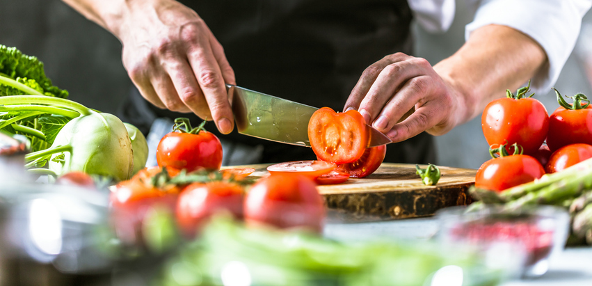 Eine Person schneidet frische Tomaten auf einem Holzbrett zwischen weiterem Gemüse.
